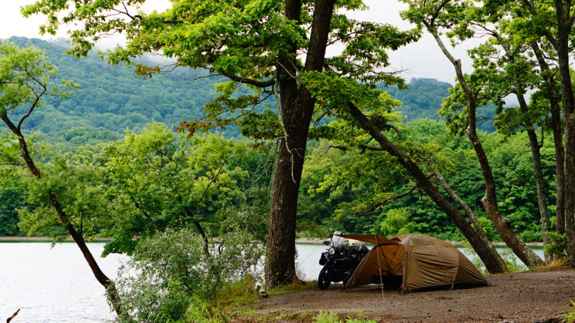十和田湖の湖畔、大きな木の下に設営されたブラウンのテント（アテナワイドツーリング130）と、その横に停められた黒のバイク（V-STROM）。背景には雨に煙る深い緑の山々と穏やかな湖が広がっている。