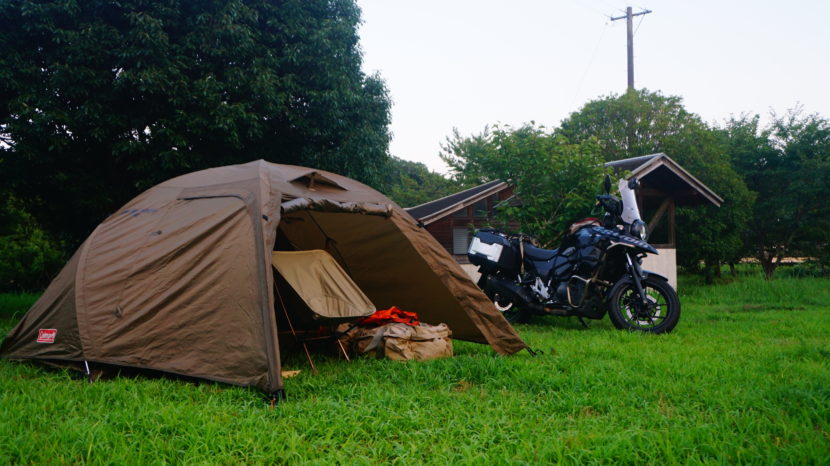 大分県の辻河原公園キャンプ場にて、芝生サイトに設営されたColemanのアテナツーリングワイド130のテントと、その横に佇むパニアケース付きのスズキ V-Strom 250。夕暮れの静かな雰囲気の中で撮影された、バイク乗り入れ可の無料キャンプ場の様子。
