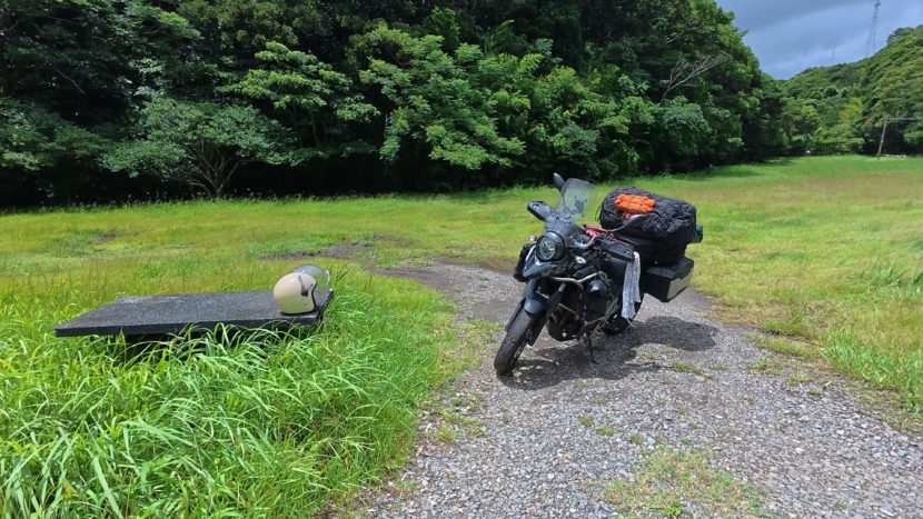 宮崎県日南市の猪崎鼻公園キャンプ場の入り口に停車する、フルパニア状態のV-STROM 250。背景には青々とした芝生と深い緑の森が広がる、九州一周ツーリング中のキャンプ場下見風景。